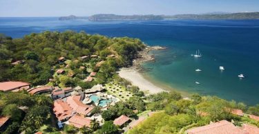 Vista aérea de un resort en Costa Rica, rodeado de exuberante vegetación tropical y con acceso directo a una playa de arena blanca con aguas cristalinas. Varias embarcaciones están ancladas cerca de la costa. El resort muestra estructuras con techos de tejas rojas, caminos bien definidos y una piscina central, ideal para turistas adultos que buscan un ambiente naturista o con ropa opcional.