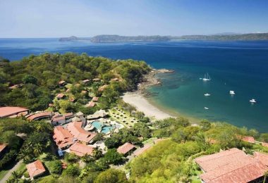 Vista aérea de un resort en Costa Rica, rodeado de exuberante vegetación tropical y con acceso directo a una playa de arena blanca con aguas cristalinas. Varias embarcaciones están ancladas cerca de la costa. El resort muestra estructuras con techos de tejas rojas, caminos bien definidos y una piscina central, ideal para turistas adultos que buscan un ambiente naturista o con ropa opcional.