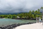 Scenic view of a cloudy beach in Kona, Hawaii, featuring turquoise waters, black volcanic rocks, and lush palm trees, offering a taste of the region's natural beauty and cultural heritage.