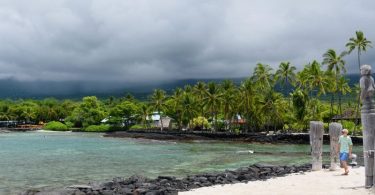 Scenic view of a cloudy beach in Kona, Hawaii, featuring turquoise waters, black volcanic rocks, and lush palm trees, offering a taste of the region's natural beauty and cultural heritage.