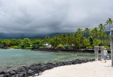 Scenic view of a cloudy beach in Kona, Hawaii, featuring turquoise waters, black volcanic rocks, and lush palm trees, offering a taste of the region's natural beauty and cultural heritage.